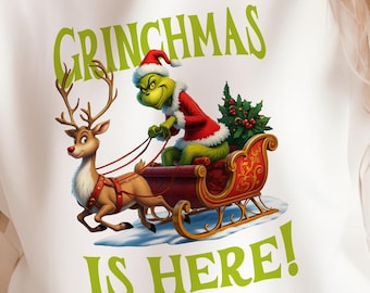 May include: White hoodie featuring a Grinch-themed design. The graphic shows the Grinch in a sleigh pulled by a reindeer, with the text "GRINCHMAS IS HERE!" in green. The design is festive and suitable for the holiday season.