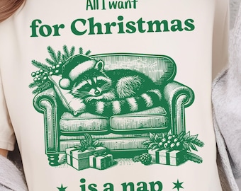 May include: A cream-coloured t-shirt featuring a green graphic of a raccoon in a Santa hat, asleep on a sofa. The text above reads "All I want for Christmas is a nap."