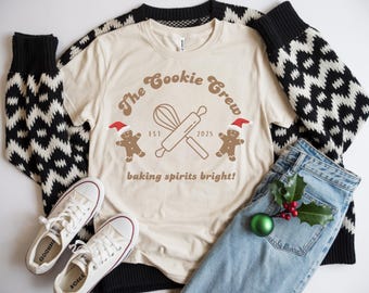 May include: A cream-colored t-shirt with the text "The Cookie Crew" and a graphic of a whisk and rolling pin. Two gingerbread men wearing Santa hats flank the design. The shirt is paired with jeans, a sweater, and white sneakers.