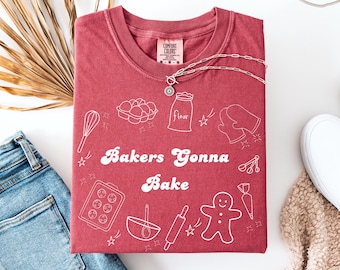 May include: A maroon Comfort Colors t-shirt with the phrase "Bakers Gonna Bake" and baking-themed illustrations in white. The shirt is folded and displayed with a silver necklace, jeans, and a white shoe.