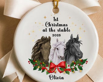 May include: A white ceramic Christmas ornament with a gold string. The ornament features three horse heads, text that reads "1st Christmas at the stable 2025", and the name "Olivia". The design includes a red bow and holly.
