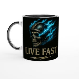 Könnte beinhalten: Weiße Keramiktasse mit schwarzem Innenraum und Griff. Die Tasse zeigt einen Totenkopf mit Motorradhelm mit blauen Flammen und den Worten "LIVE FAST" in goldener Schrift.