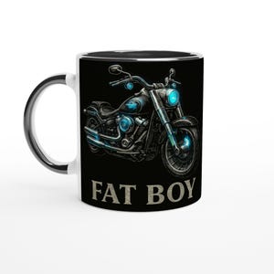 Può includere: Tazza in ceramica bianca con interno e manico neri. La tazza presenta un'illustrazione dettagliata di una motocicletta con dettagli blu e la scritta "FAT BOY" in grassetto. Il design della moto è centrato sulla tazza.