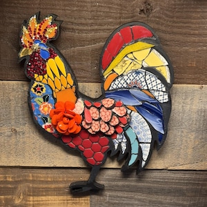 May include: A colorful mosaic rooster wall art piece. The rooster features a black outline with a vibrant mosaic design. The mosaic incorporates red, orange, yellow, blue, and white tiles, creating a textured and eye-catching artwork. The rooster is mounted on a wooden background.
