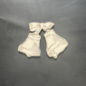 May include: Cream-colored decorative bells with gold trim and a bow. The bells are connected by a bow at the top. The bells have a detailed design with lines. The background is a dark gray color.