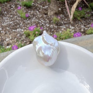 Puede incluir: Una gran perla barroca iridiscente con brillo nacarado descansa en un cuenco de cer&aacute;mica blanco. La superficie de la perla refleja un espectro de colores. El fondo presenta vegetaci&oacute;n y flores moradas.
