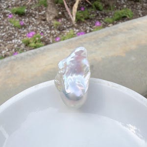 Puede incluir: Una gran perla barroca iridiscente descansa en un cuenco blanco poco profundo. La perla tiene una forma irregular y lisa con una superficie brillante que refleja un espectro de colores. El cuenco est&aacute; sobre un fondo exterior borroso.