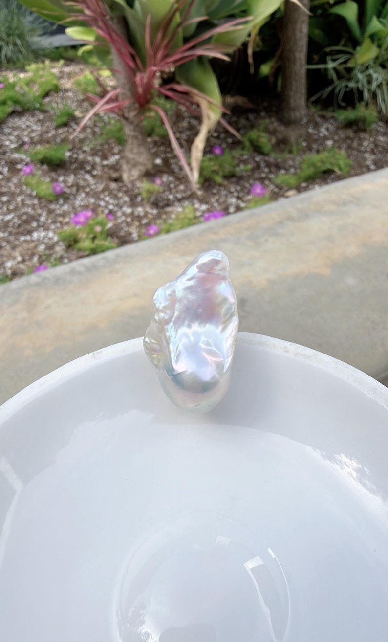 Puede incluir: Una gran perla barroca iridiscente descansa en un cuenco blanco poco profundo. La perla tiene una forma irregular y lisa con una superficie brillante que refleja un espectro de colores. El cuenco est&aacute; sobre un fondo exterior borroso.