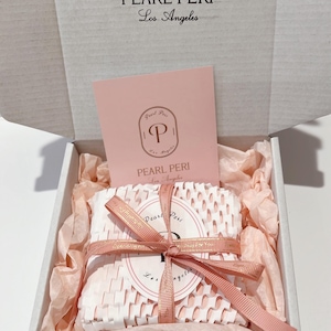 May include: A gift box with the text "PEARL PERI Los Angeles" on the lid. Inside, a wrapped item is secured with a pink ribbon. The box contains pink tissue paper and a pink card with the brand's logo.