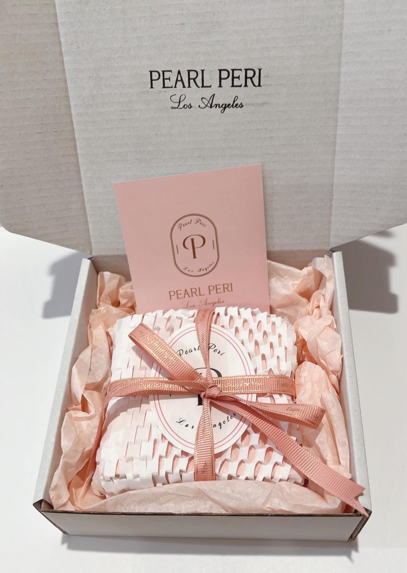 May include: A gift box with the text "PEARL PERI Los Angeles" on the lid. Inside, a wrapped item is secured with a pink ribbon. The box contains pink tissue paper and a pink card with the brand's logo.