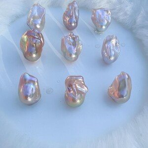 May include: Nine iridescent baroque pearls with a mix of pink, gold, and lavender hues. The pearls have an irregular shape and a lustrous finish. They are arranged on a white surface, set against a soft, white, fluffy background.