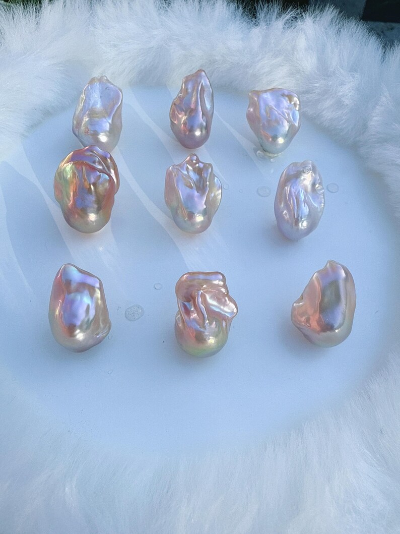 May include: Nine iridescent baroque pearls with a mix of pink, gold, and lavender hues. The pearls have an irregular shape and a lustrous finish. They are arranged on a white surface, set against a soft, white, fluffy background.