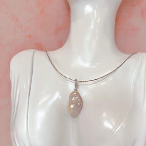 May include: A white bust displays a silver necklace with a large, iridescent baroque pearl pendant. The pearl has a unique, irregular shape and a soft, pearlescent sheen. The background is a light pink.