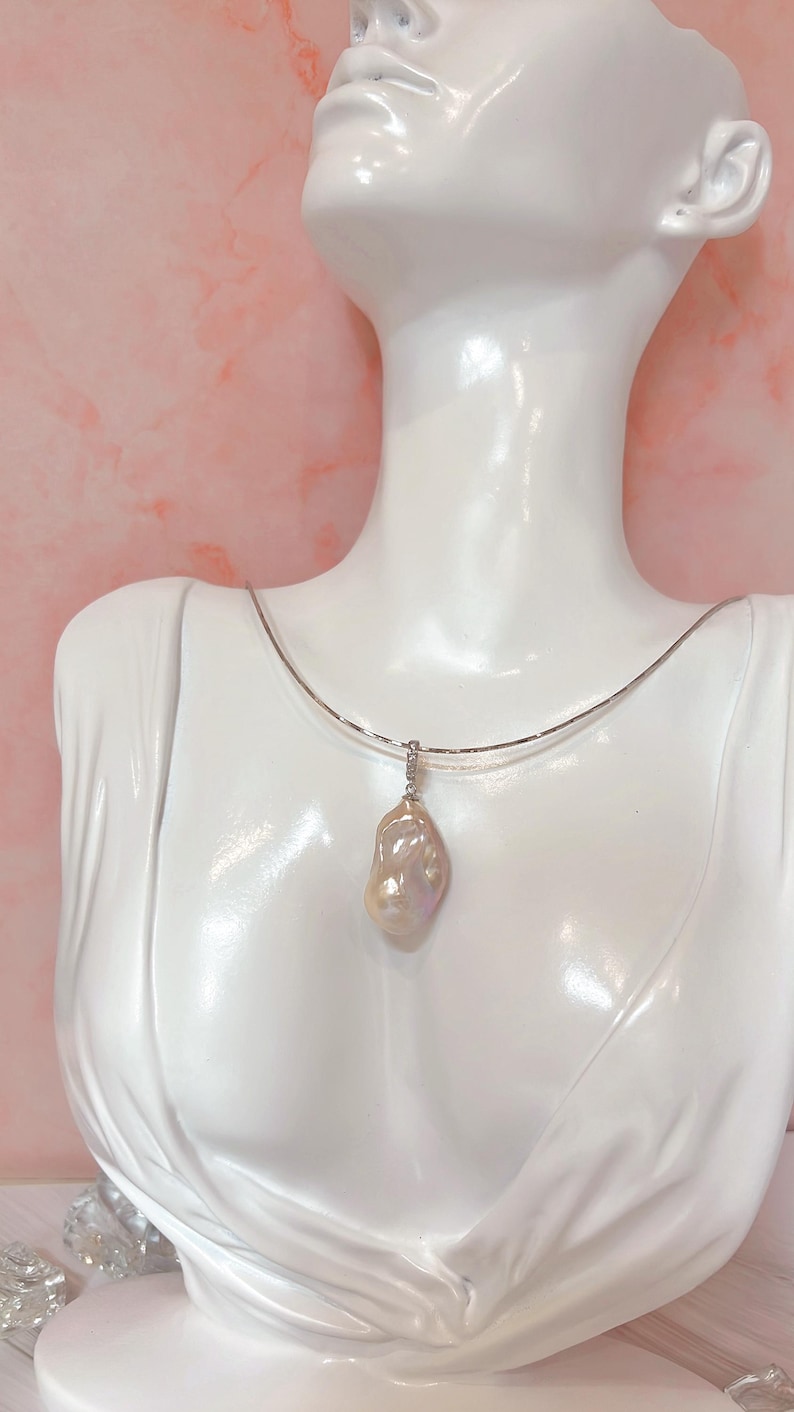 May include: A white bust displays a silver necklace with a large, iridescent baroque pearl pendant. The pearl has a unique, irregular shape and a soft, pearlescent sheen. The background is a light pink.