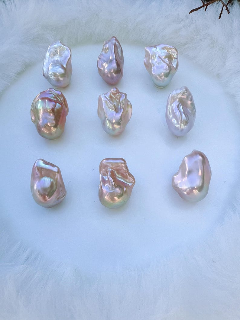 May include: Nine iridescent baroque pearls with a lustrous, pearlescent sheen. The pearls display a range of colours, including pink, gold, and white, and are arranged on a white, fluffy surface. The irregular shapes of the pearls add to their unique appeal.