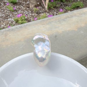 Puede incluir: Una gran perla barroca iridiscente descansa en un cuenco de cer&aacute;mica blanco. La perla tiene una forma irregular y lisa con una superficie brillante que refleja un espectro de colores. El cuenco es redondo y tiene un acabado brillante.