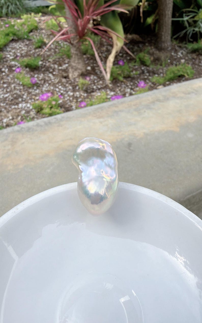 Puede incluir: Una gran perla barroca iridiscente descansa en un cuenco de cer&aacute;mica blanco. La perla tiene una forma irregular y lisa con una superficie brillante que refleja un espectro de colores. El cuenco es redondo y tiene un acabado brillante.