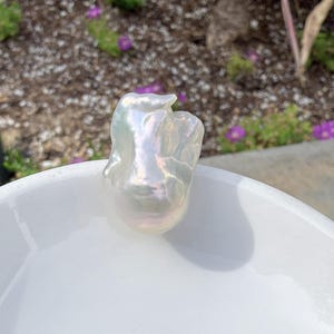 Puede incluir: Una perla grande, de forma irregular e iridiscente, con una superficie lisa y brillante. La perla est&aacute; en un cuenco blanco, reflejando la luz y mostrando su forma natural y org&aacute;nica. Los colores de la perla cambian del blanco a tonos sutiles.