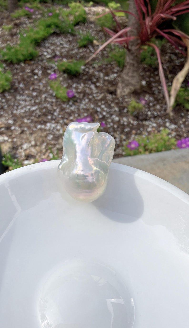 Puede incluir: Una perla grande, de forma irregular e iridiscente, con una superficie lisa y brillante. La perla est&aacute; en un cuenco blanco, reflejando la luz y mostrando su forma natural y org&aacute;nica. Los colores de la perla cambian del blanco a tonos sutiles.