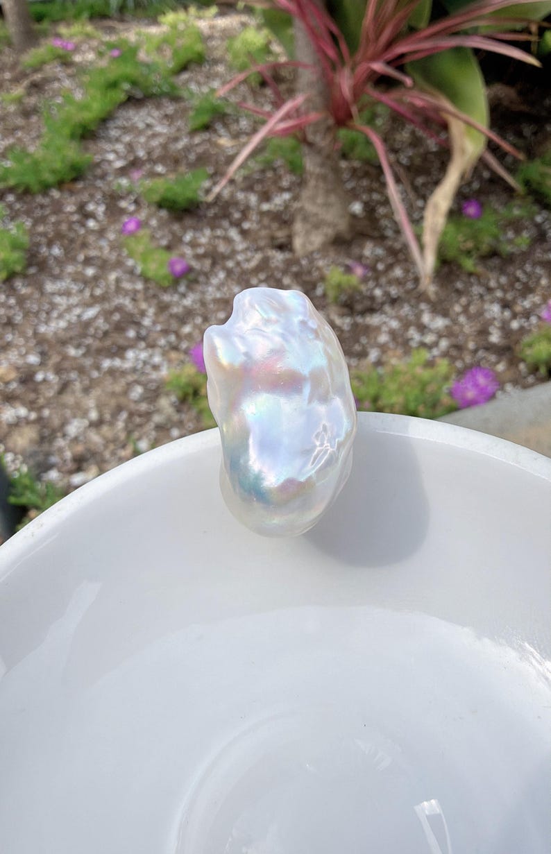 Puede incluir: Una perla blanca grande, de forma irregular, con colores iridiscentes. La perla se encuentra sobre una superficie de cer&aacute;mica blanca, con un fondo borroso de plantas verdes y flores moradas.