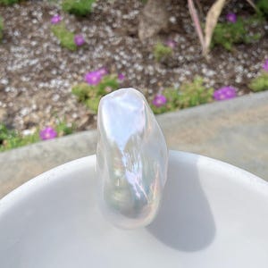 Puede incluir: Una gran perla barroca iridiscente descansa en un cuenco de cer&aacute;mica blanco. La perla tiene una forma irregular y lisa con una superficie brillante que refleja la luz, mostrando una gama de colores. El cuenco est&aacute; al aire libre.