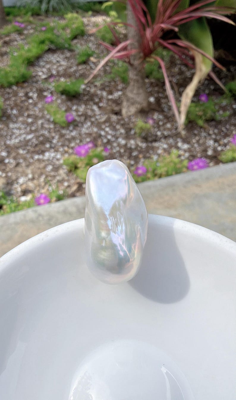 Puede incluir: Una gran perla barroca iridiscente descansa en un cuenco de cer&aacute;mica blanco. La perla tiene una forma irregular y lisa con una superficie brillante que refleja la luz, mostrando una gama de colores. El cuenco est&aacute; al aire libre.