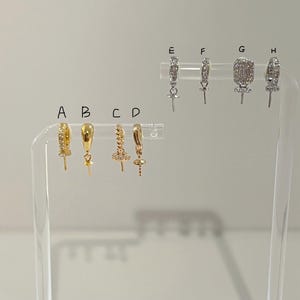 May include: A selection of gold and silver earrings on a clear display stand. The earrings showcase diverse designs, including hoops, studs, and drop styles. Some are embellished with gemstones, while others have detailed metalwork. The earrings are set against a plain backdrop.