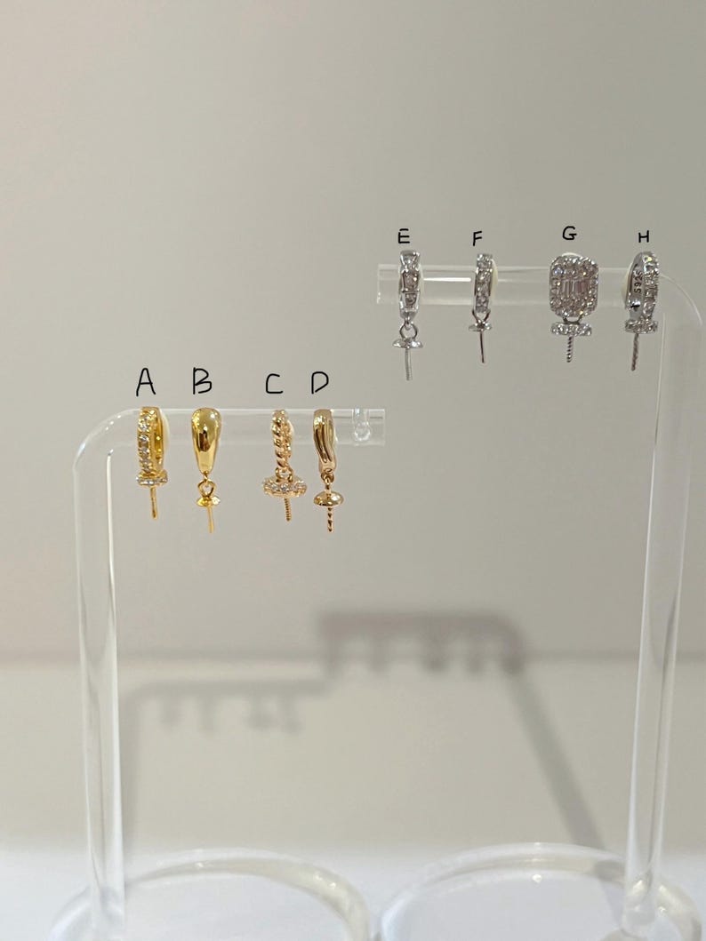 May include: A selection of gold and silver earrings on a clear display stand. The earrings showcase diverse designs, including hoops, studs, and drop styles. Some are embellished with gemstones, while others have detailed metalwork. The earrings are set against a plain backdrop.