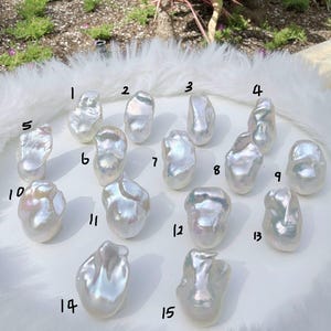 Puede incluir: Quince perlas blancas irregulares y brillantes se exhiben sobre una superficie blanca y esponjosa. Cada perla tiene una forma org&aacute;nica &uacute;nica, que refleja la luz con tonos iridiscentes. Las perlas est&aacute;n numeradas del 1 al 15.