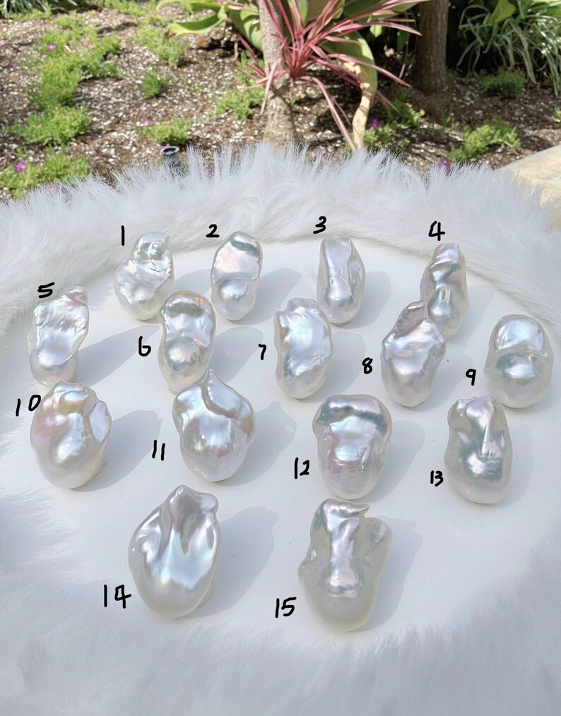 Puede incluir: Quince perlas blancas irregulares y brillantes se exhiben sobre una superficie blanca y esponjosa. Cada perla tiene una forma org&aacute;nica &uacute;nica, que refleja la luz con tonos iridiscentes. Las perlas est&aacute;n numeradas del 1 al 15.