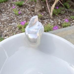 Puede incluir: Una perla blanca grande, lustrosa y de forma irregular, se encuentra sobre un cuenco de cer&aacute;mica blanco y brillante. La perla tiene un brillo nacarado, que refleja la luz. El fondo presenta vegetaci&oacute;n y flores moradas, a&ntilde;adiendo contraste natural.