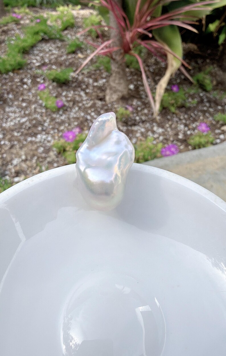 Puede incluir: Una perla blanca grande, lustrosa y de forma irregular, se encuentra sobre un cuenco de cer&aacute;mica blanco y brillante. La perla tiene un brillo nacarado, que refleja la luz. El fondo presenta vegetaci&oacute;n y flores moradas, a&ntilde;adiendo contraste natural.