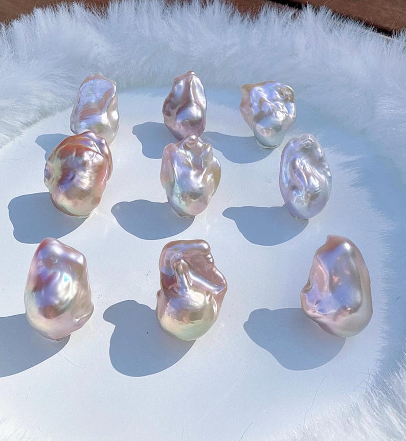 May include: Nine iridescent baroque pearls with a lustrous, pearlescent sheen. The pearls display a range of colours, including pink, peach, and white, and are irregularly shaped. They are arranged on a white surface.