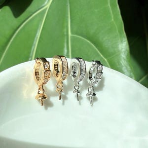 Puede incluir: Cuatro pendientes de aro en tonos dorados y plateados, cada uno con una peque&ntilde;a clavija colgante. Los pendientes est&aacute;n adornados con peque&ntilde;as piedras preciosas transparentes. Los pendientes se exhiben sobre una superficie blanca y curvada sobre un fondo verde.
