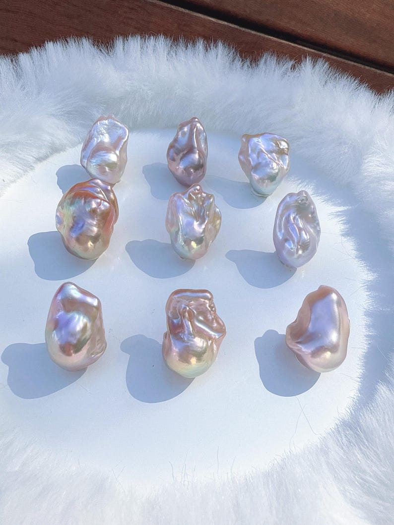 May include: Nine irregularly shaped baroque pearls with a shimmering, iridescent surface. The pearls exhibit a spectrum of colours, including pink, lavender, and gold, presented on a white surface. Each pearl has a unique form and size.