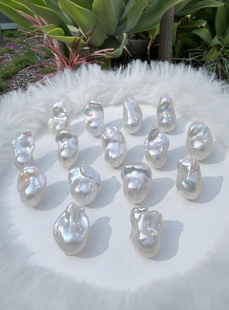 Puede incluir: Una colecci&oacute;n de perlas blancas brillantes de forma irregular. Las perlas est&aacute;n dispuestas sobre una superficie blanca con una textura suave y esponjosa. Las perlas tienen un brillo nacarado, que refleja la luz y crea una sutil iridiscencia. El tema general es el lujo y la belleza natural.