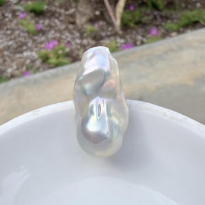Puede incluir: Una gran perla barroca iridiscente con un brillo nacarado. La forma irregular de la perla se muestra sobre un fondo blanco. La perla refleja la luz, mostrando una gama de colores. Esta perla &uacute;nica es una pieza llamativa.