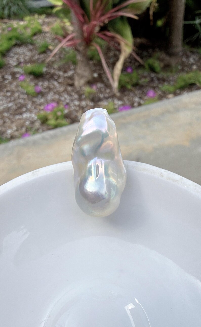 Puede incluir: Una gran perla barroca iridiscente con un brillo nacarado. La forma irregular de la perla se muestra sobre un fondo blanco. La perla refleja la luz, mostrando una gama de colores. Esta perla &uacute;nica es una pieza llamativa.