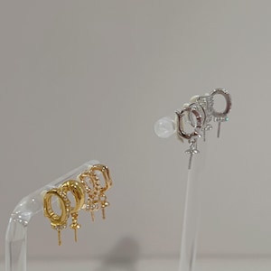 May include: A selection of gold and silver hoop earrings. The gold earrings feature different designs, some with small clear stones and hanging details. The silver earrings also have clear stones and a cross pendant. All earrings are displayed on clear stands.