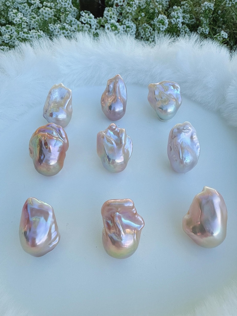 May include: Nine iridescent baroque pearls with a pearlescent sheen. The pearls display pink, gold, and silver hues, and are arranged on a white, fluffy surface. The pearls have irregular shapes, adding to their unique appeal. These are ideal for jewellery making.