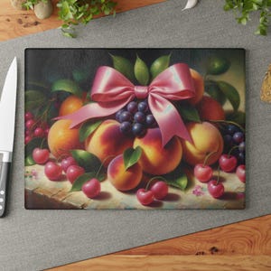 Könnte beinhalten: Ein Schneidebrett aus Glas mit einem Stillleben aus Obst und einer rosafarbenen Schleife. Das Obst umfasst Pfirsiche, Kirschen und Trauben.