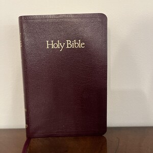 Könnte beinhalten: Eine weinrote Heilige Bibel mit goldener Schrift. Das Buch ist geschlossen und steht aufrecht, mit einem Leseband. Der Einband hat eine glatte, lederartige Textur.