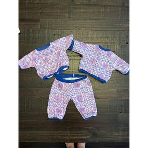 Könnte beinhalten: Ein Vintage-Puppenkleidungsset mit zwei kurzärmeligen Oberteilen und einer Hose. Das Set ist mit einem sich wiederholenden Muster aus rosa und blauen Quadraten auf weißem Grund mit blauen Verzierungen versehen. Die Kleidung ist wahrscheinlich für eine Puppe.