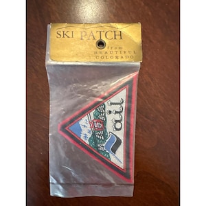 May include: A triangular embroidered ski patch featuring a mountain landscape with a ski lift and the text Vail. The patch is enclosed in a clear plastic sleeve with a gold cardboard header that reads SKI PATCH from BEAUTIFUL COLORADO.