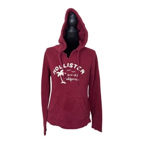 May include: A maroon long-sleeve hooded sweatshirt featuring a white graphic print on the chest that reads Hollister DIV-22 California with a palm tree icon. The pullover includes a front kangaroo pocket and matching drawstring cords.