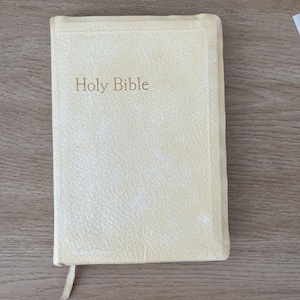 May include: A cream-coloured Holy Bible with the title embossed on the cover. The book has a textured surface and a cream-coloured ribbon bookmark. The book is closed and resting on a wooden surface.