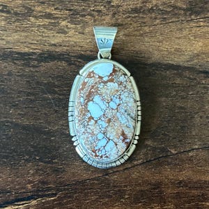 May include: An oval pendant featuring a white and brown matrix stone set in a polished silver frame. The silver border displays etched vertical lines along the sides and bottom, and the bail at the top includes a small stamped sunburst design.