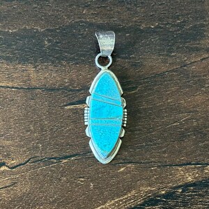 May include: A marquise-shaped pendant featuring inlaid turquoise stone segments separated by thin silver lines. The pendant has a polished silver frame with a notched edge and a silver bail at the top for attachment to a chain or cord.