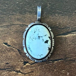 May include: An oval pendant featuring a pale, light blue stone with dark matrix veining, set in a scalloped silver frame with etched line details along the border and a vertical silver bail at the top for attachment to a chain or cord.