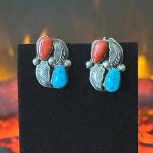 May include: A pair of silver earrings featuring a teardrop-shaped red coral stone and a teardrop-shaped blue turquoise stone. Each earring is accented with silver leaf motifs and small silver spheres, creating a symmetrical design with a polished finish.